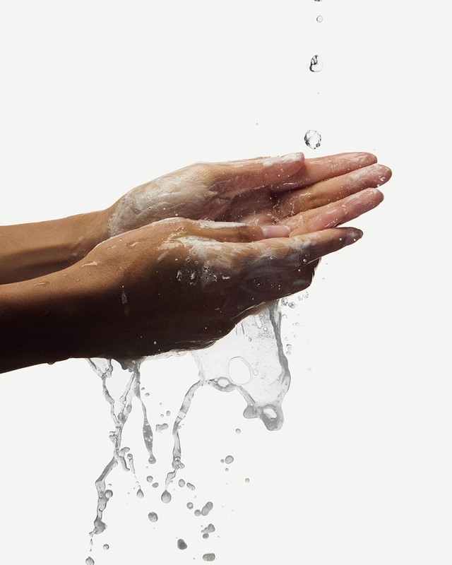 Woman's hands with soap under running water, washing hands against a plain white background in a beauty or healthcare context.