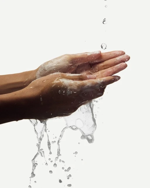 Woman's hands with soap under running water, washing hands against a plain white background in a beauty or healthcare context.
