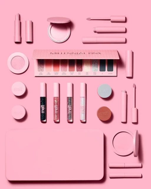 Flat lay of Millennial Pink eyeshadow palette surrounded by pink makeup products, including lipstick, glosses, and compacts, on a pink background.