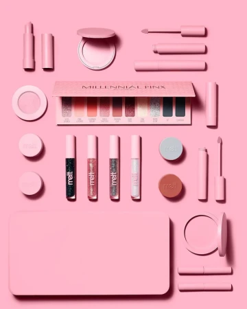 Flat lay of Millennial Pink eyeshadow palette surrounded by pink makeup products, including lipstick, glosses, and compacts, on a pink background.