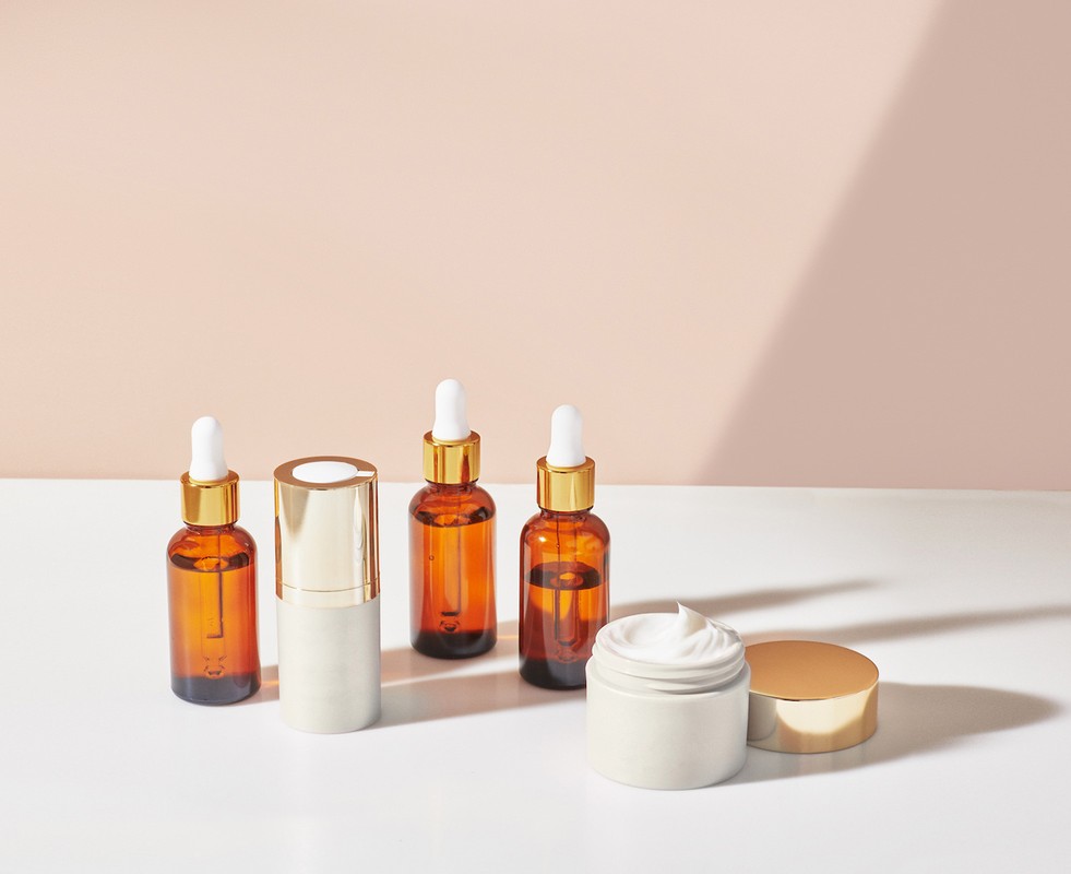 Cosmetic product bottles and jar on white surface with a light peach background and shadows. Brown glass and gold details.