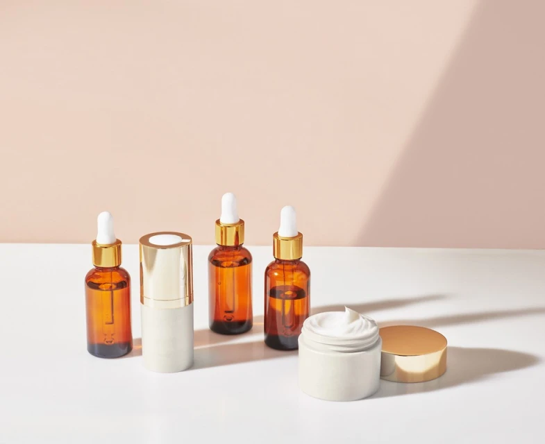 Cosmetic product bottles and jar on white surface with a light peach background and shadows. Brown glass and gold details.