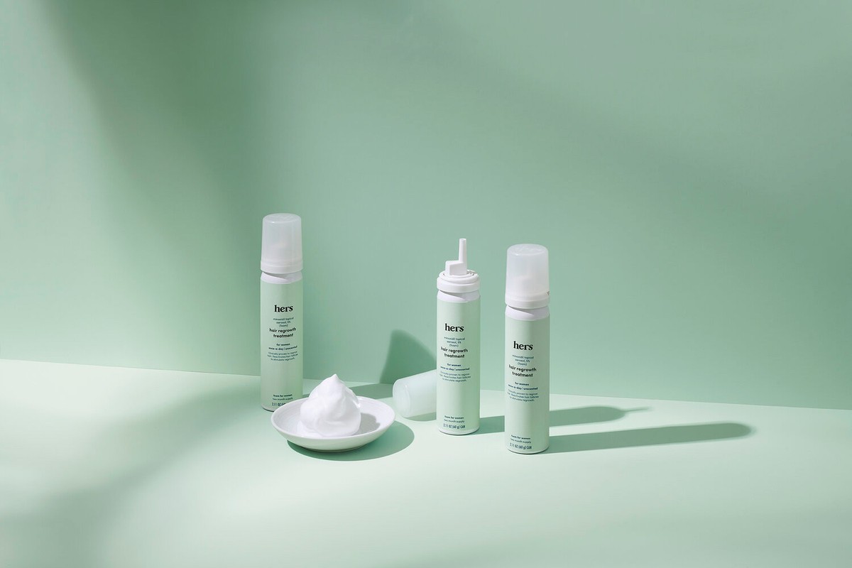 Product shot of three 'hers' hair regrowth treatment cans with a dollop of foam on a plate, set against a light green backdrop.