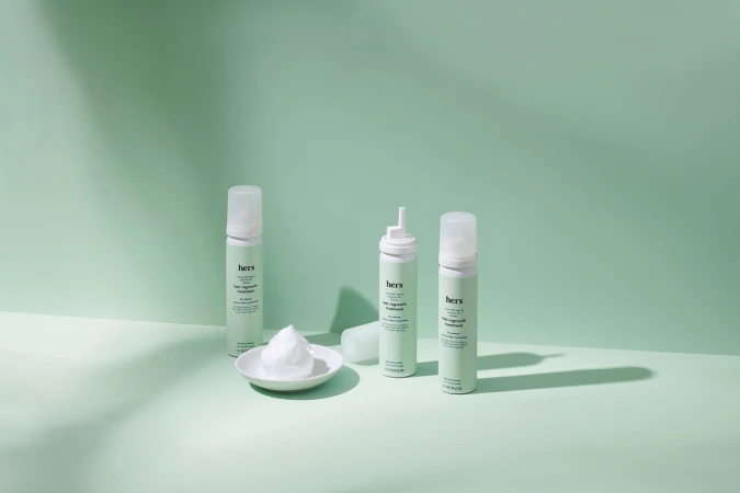Product shot of three 'hers' hair regrowth treatment cans with a dollop of foam on a plate, set against a light green backdrop.