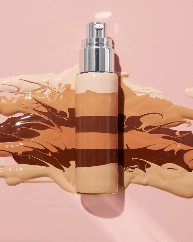 Still life shot of foundation bottle with pump surrounded by swirling strokes of liquid foundation on light pink background.