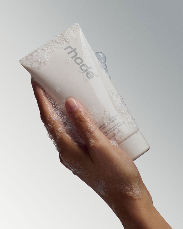 Hand covered in soap suds holding a white skincare tube against a grey studio backdrop