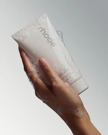 Hand covered in soap suds holding a white skincare tube against a grey studio backdrop