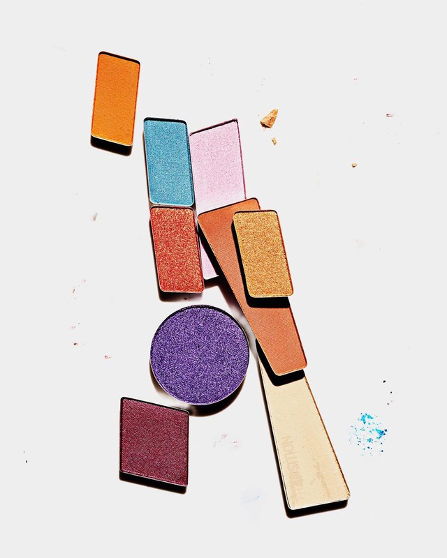 Overhead shot of arranged eyeshadow palettes in various shapes and colors, including orange, purple, and gold, on a white surface.