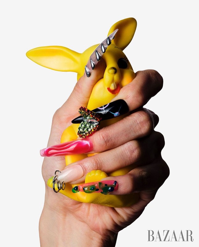 Hand holding a yellow toy rabbit with long, decorative, and colorful fingernails featuring jewels and various designs.