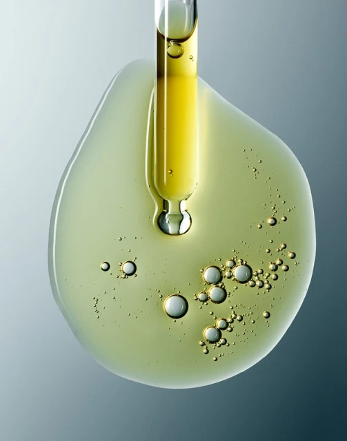 Close-up of a dropper dispensing yellow oil onto a surface, creating a small pool with visible bubbles against a grey background.