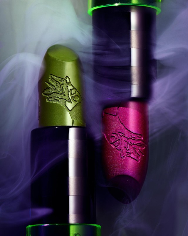 Close-up studio shot of green and purple lipsticks with embossed designs against a blurred purple and white smoky background.