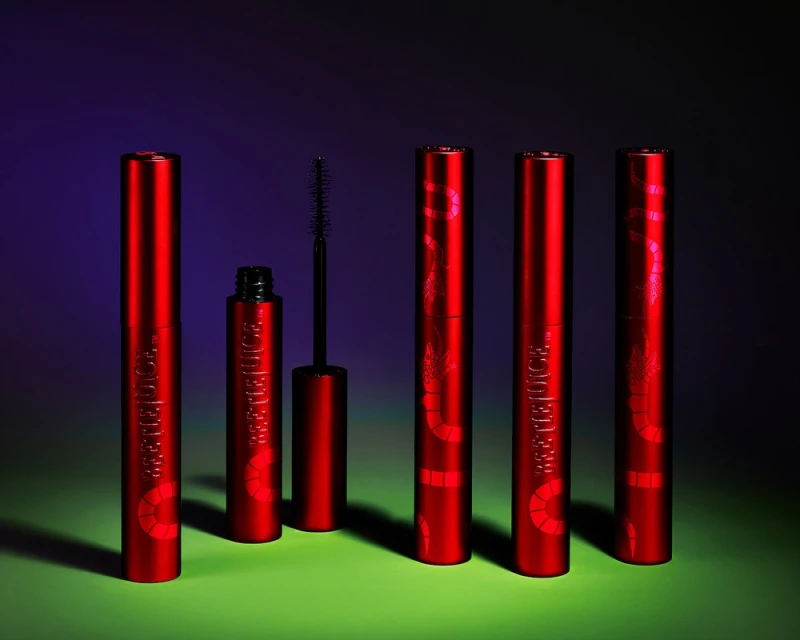 Red tubes of Beetlejuice-themed mascara are presented in studio lighting against a gradient background, one is open with the brush visible.