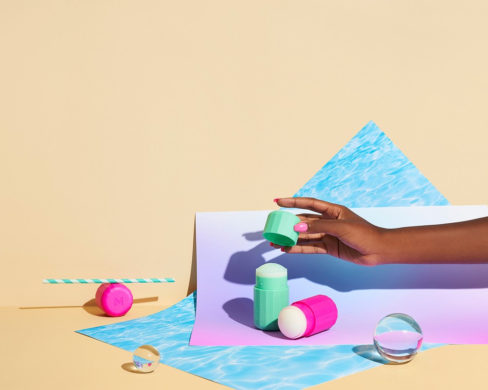 Cosmetic product display featuring female hand holding a green container, with geometric shapes and pastel colors on a light backdrop.