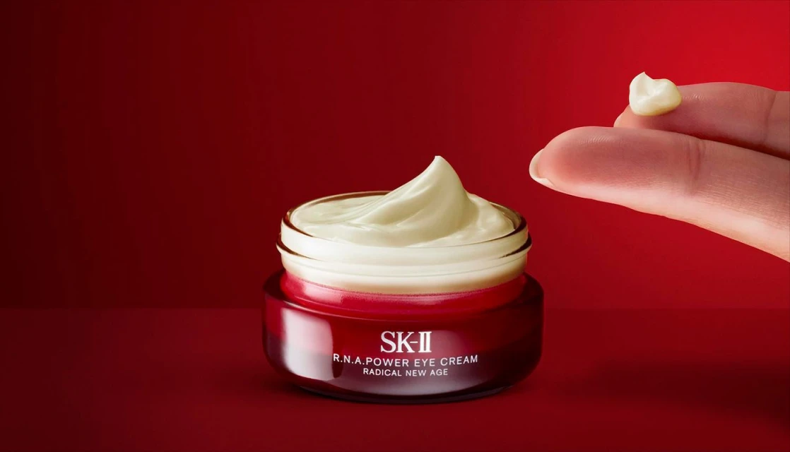 Close-up of SK-II R.N.A. Power Eye Cream jar with finger holding a dollop of cream on a red background. Beauty product shot.