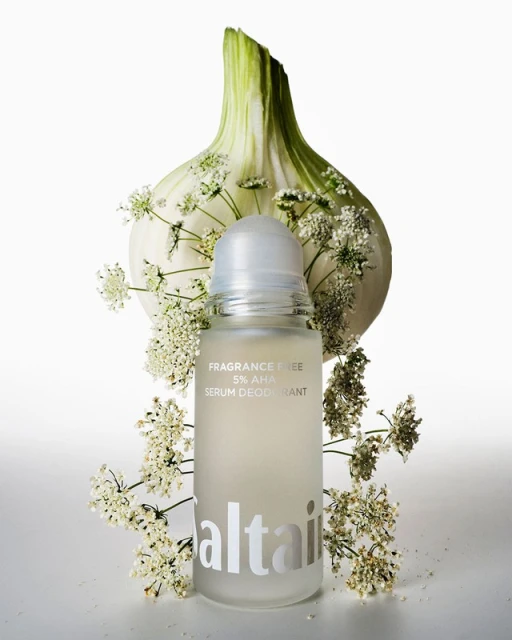Product shot of Altair serum deodorant with fennel and white flowers. White background, clean aesthetic, focus on product and natural elements.