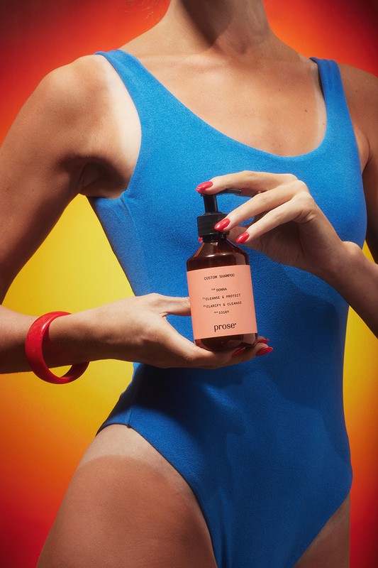 Woman in blue bathing suit holds prose brand shampoo bottle against an orange gradient background with a red bracelet on wrist.