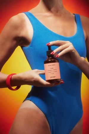 Woman in blue bathing suit holds prose brand shampoo bottle against an orange gradient background with a red bracelet on wrist.