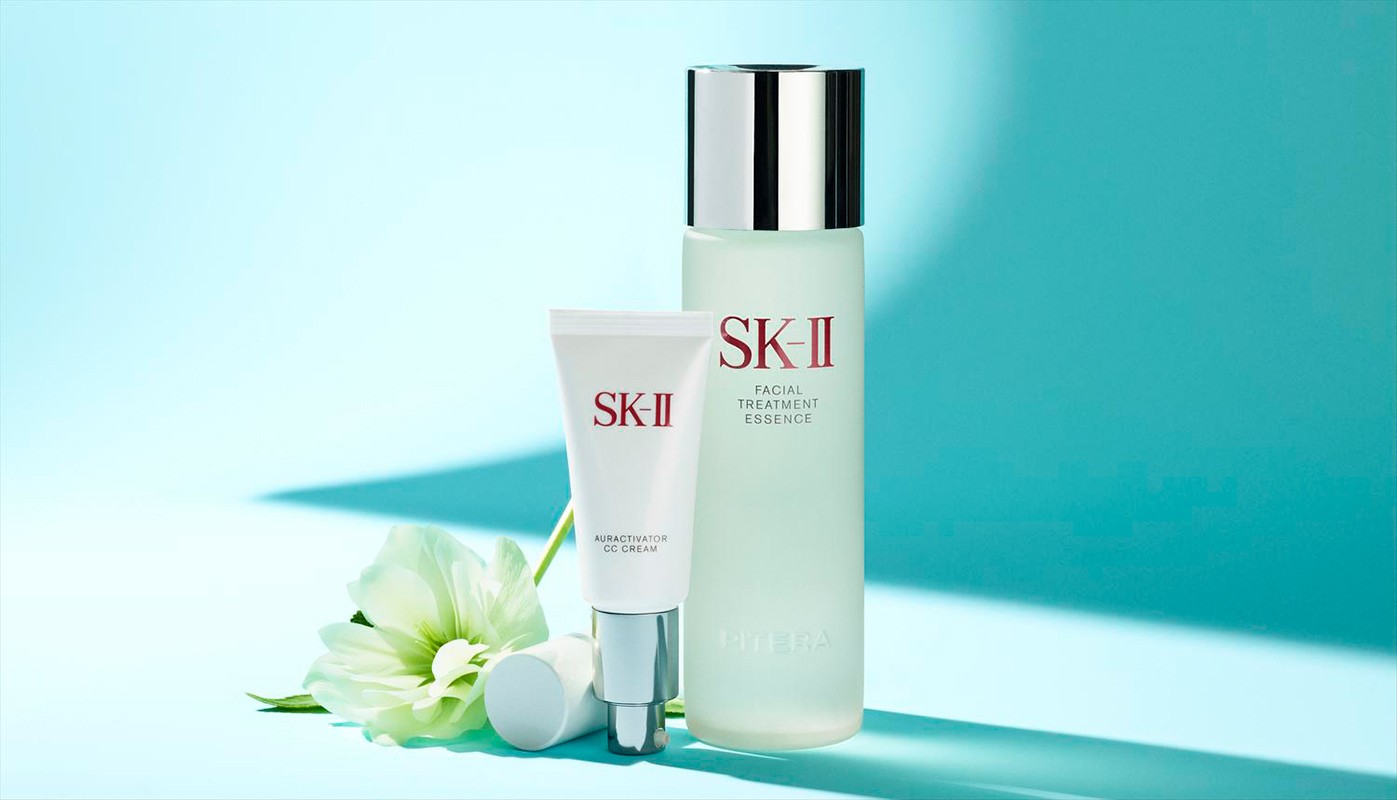 Still life shot of SK-II skincare products including a facial treatment essence, a CC cream, and a white flower against a light blue background.