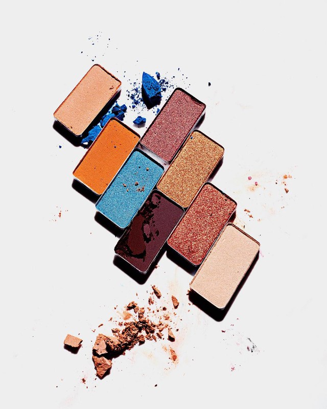Eye shadow palette with multiple colors including brown, blue, orange, and red on a white surface, with some powder scattered.