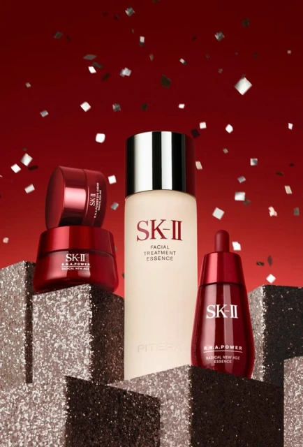 Elevated view of SK-II skincare products: a bottle of facial treatment essence, eye cream, and a serum against a red glitter background.