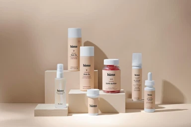 Collection of Hims hair care product bottles and jars arranged on tiered beige blocks against a beige backdrop.