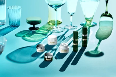 Skincare product jars and bottles arranged on a bright blue surface among colourful glassware casting long shadows.