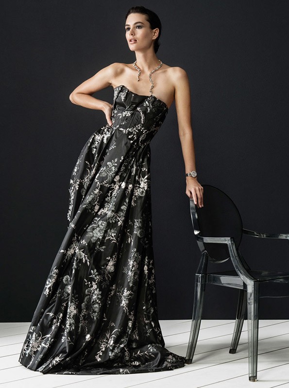 Model in black floral gown leaning on a transparent acrylic chair against a dark studio background