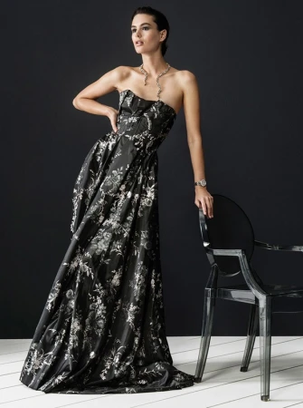 Model in black floral gown leaning on a transparent acrylic chair against a dark studio background