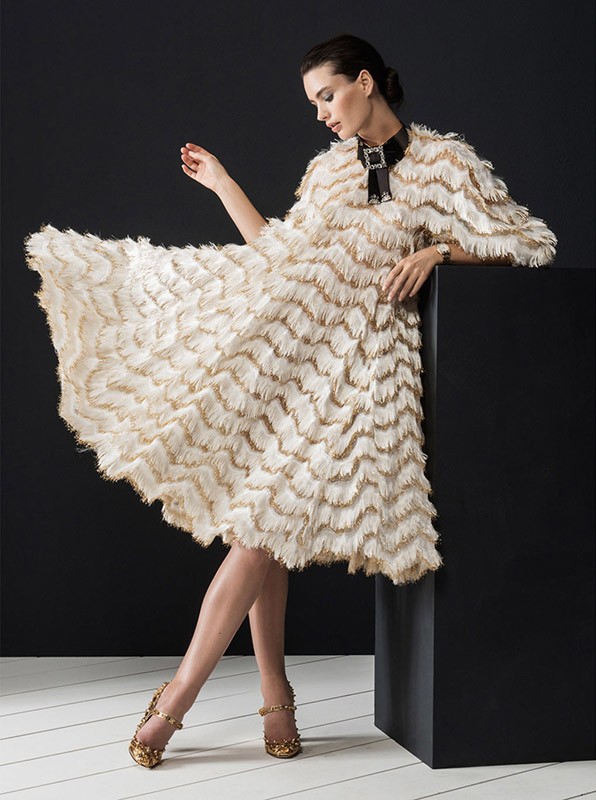 Woman in feathered dress poses next to black cube in photo studio wearing jeweled heels and a dark choker, against a dark background.