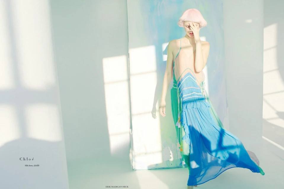 Woman with pink hair wears a long blue ombre dress in a brightly lit studio, fashion editorial.