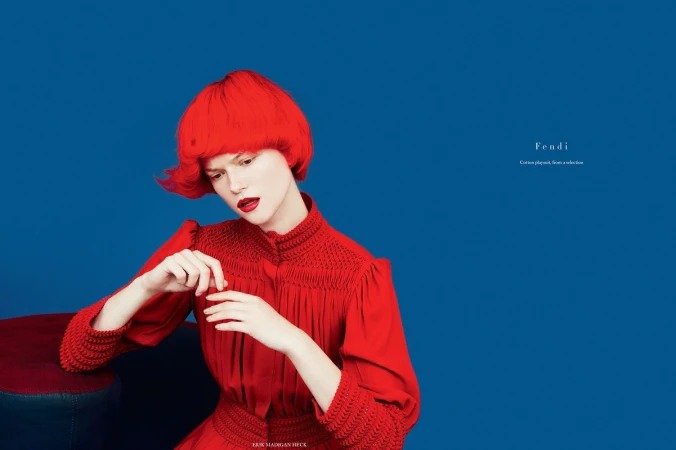 Model with bright red hair wearing a red high-necked dress against a solid blue background by Andrea Huelse