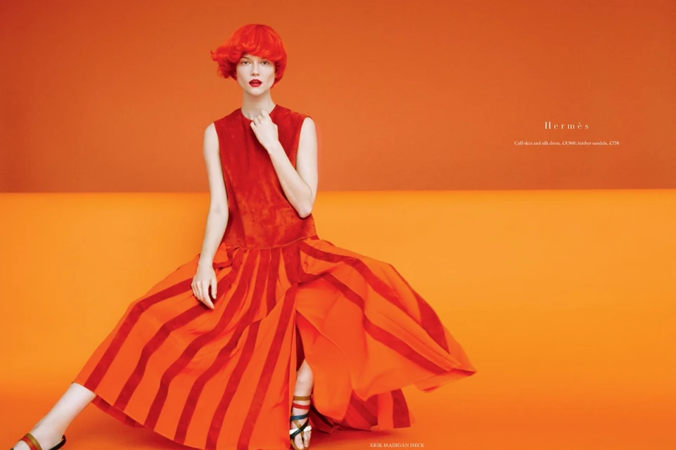 Fashion editorial shot: A model with an orange wig poses in an orange and red dress against an orange studio backdrop with text overlay.