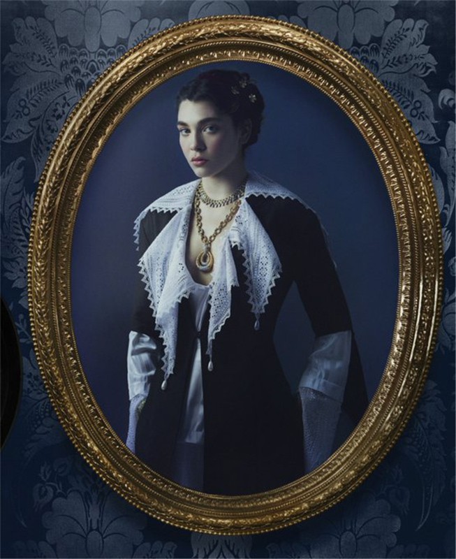 Portrait of a young woman in period clothing and gold jewelry, framed in an ornate gold oval frame against a blue patterned wall.