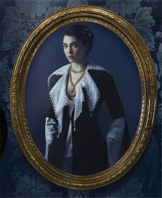 Portrait of a young woman in period clothing and gold jewelry, framed in an ornate gold oval frame against a blue patterned wall.