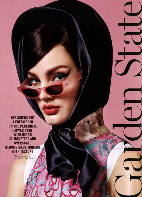 Fashion editorial shot of a woman with winged eyeliner, sunglasses, and a black headscarf, set against a pale pink background.