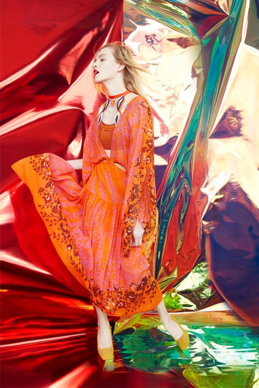 A woman models a flowing floral dress with long sleeves against a vibrant, textured backdrop of red and iridescent panels.