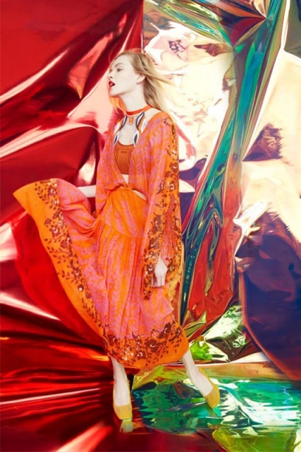 A woman models a flowing floral dress with long sleeves against a vibrant, textured backdrop of red and iridescent panels.