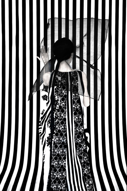 Woman with back turned wearing a black and white patterned dress in front of a black and white striped backdrop with a transparent fabric.