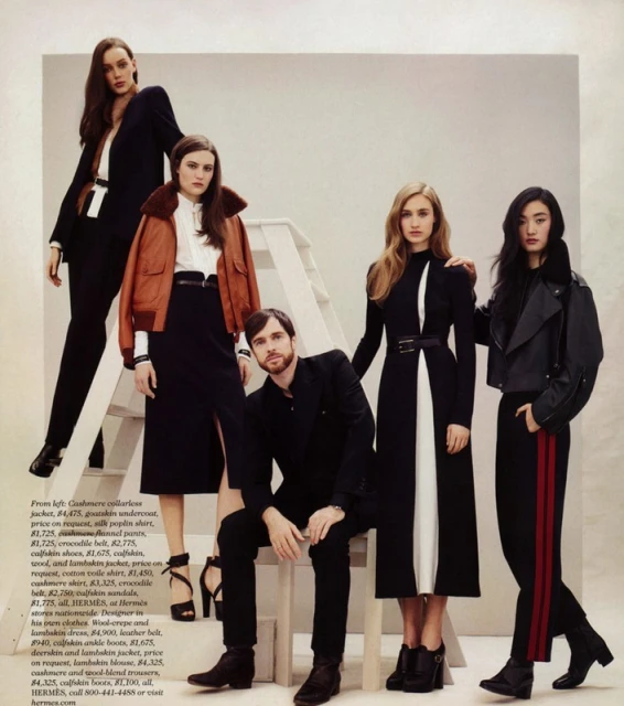 Five fashion models showcase designer apparel indoors; 4 women and 1 man posing. One woman stands on a ladder.