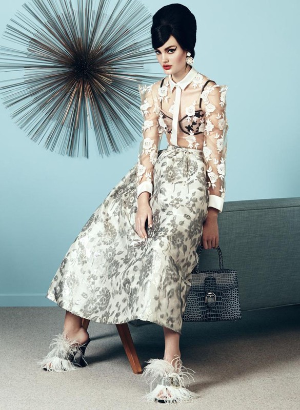 A fashion shot of a poised adult female with a bouffant hairstyle, in a floral skirt, sheer blouse, and feathered heels, seated indoors.