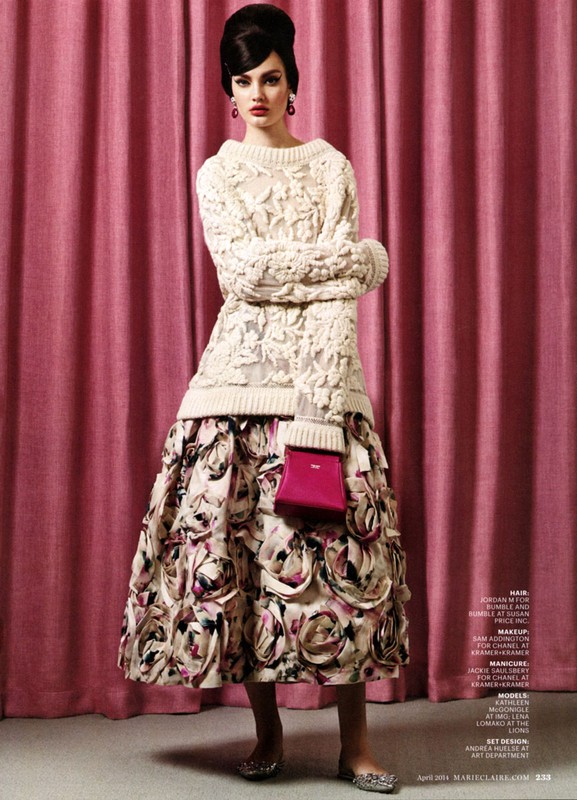 A woman poses in an off-white knit sweater and floral skirt, holding a bright pink purse, against a pink curtain backdrop in a fashion shot.