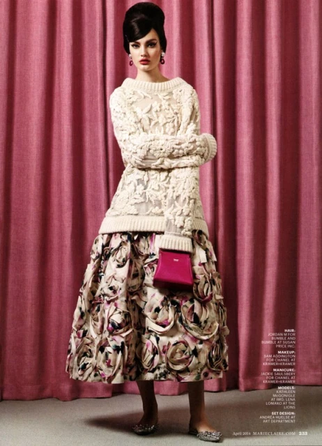 A woman poses in an off-white knit sweater and floral skirt, holding a bright pink purse, against a pink curtain backdrop in a fashion shot.