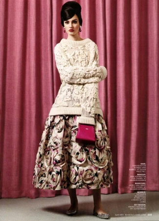 A woman poses in an off-white knit sweater and floral skirt, holding a bright pink purse, against a pink curtain backdrop in a fashion shot.