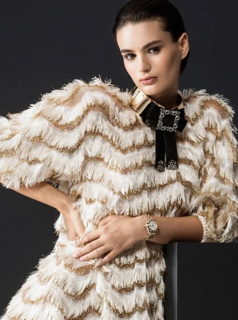 Model in white and gold fringed dress with black bow tie and metallic choker against dark background