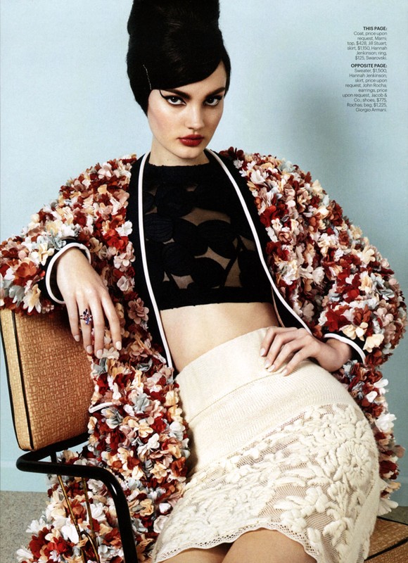 Model with dark retro updo seated on a chair wearing a heavily textured floral coat over a black crop top and cream skirt.
