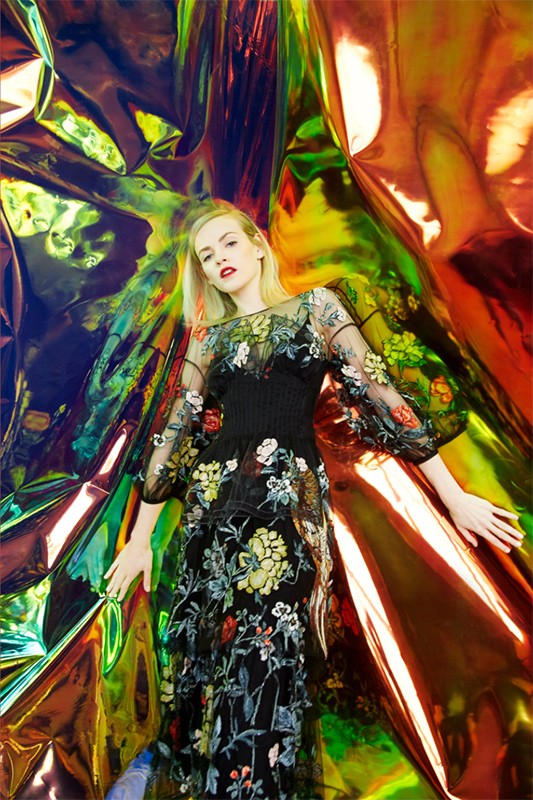 Woman with light blonde hair posing in a black dress with floral embroidery against a shiny, reflective, iridescent backdrop.