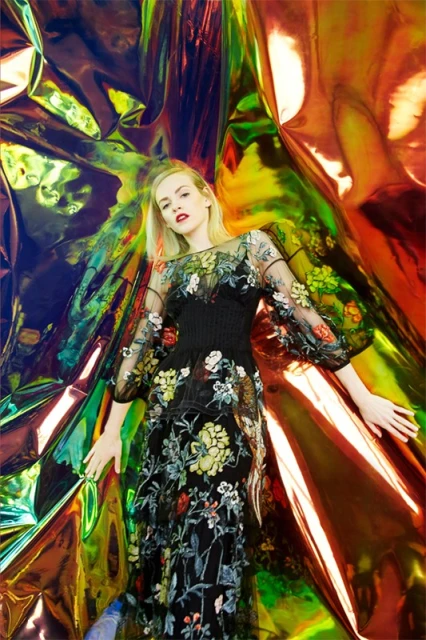 Woman with light blonde hair posing in a black dress with floral embroidery against a shiny, reflective, iridescent backdrop.