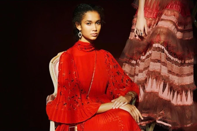 A dark-haired woman in a red dress sits in an ornate chair, looking into the camera with a neutral expression in a studio setting.