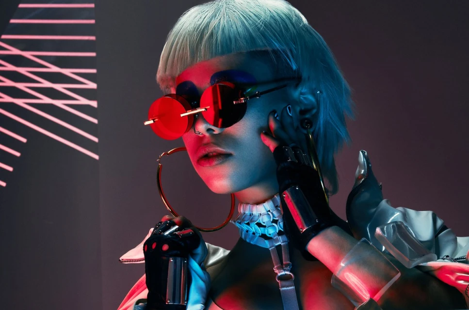 Close-up of a person with short blue hair, wearing round red glasses and a futuristic outfit, posing in a studio with neon lighting.