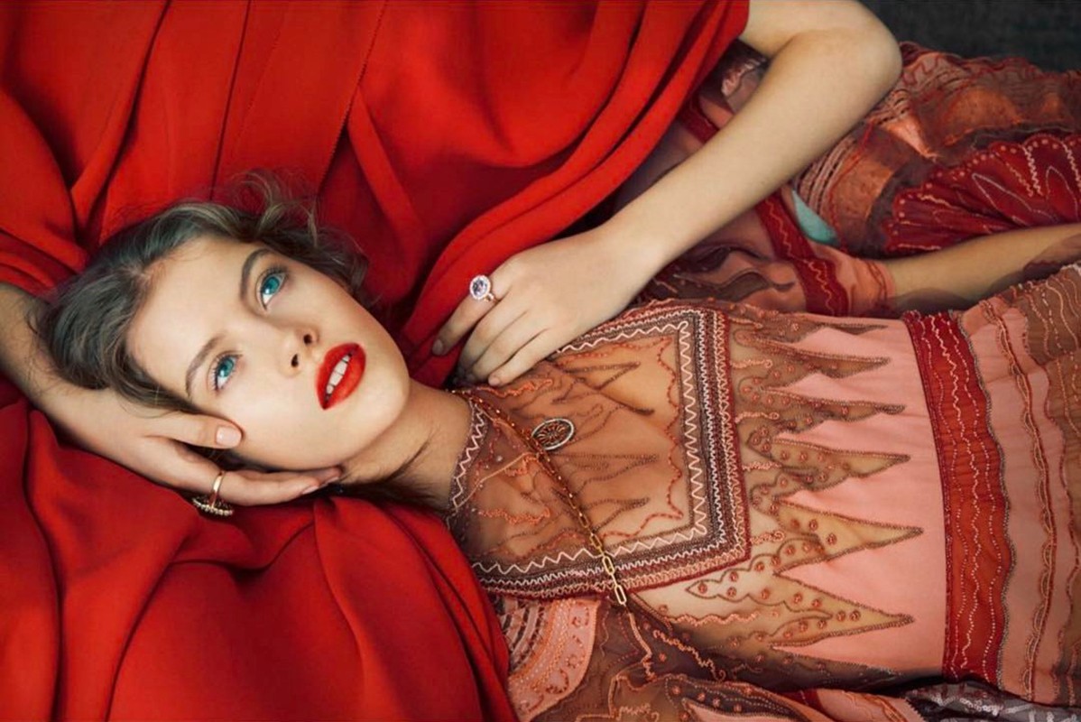 Close-up portrait of a woman with red lips and blue eyes lying amidst draped red fabric and patterned garments designed by Andrea Huelse.